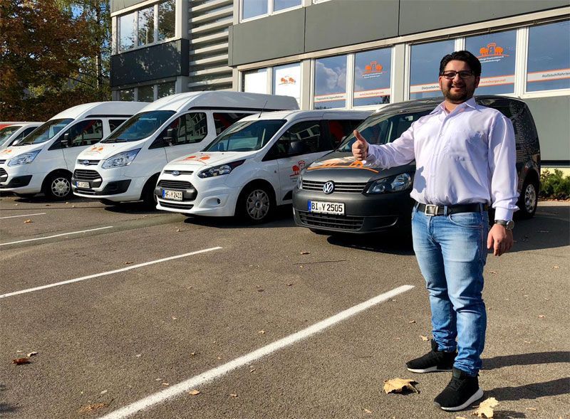 Ein Mann mit Daumen hoch steht vor einer Reihe von Vans und lächelt in die Kamera